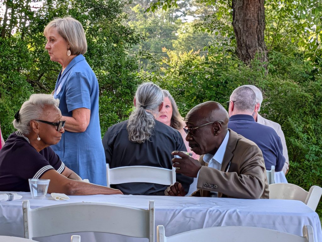 People sitting and conversing at outdoor event with white chairs and tables, surrounded by green trees and foliage in the background.