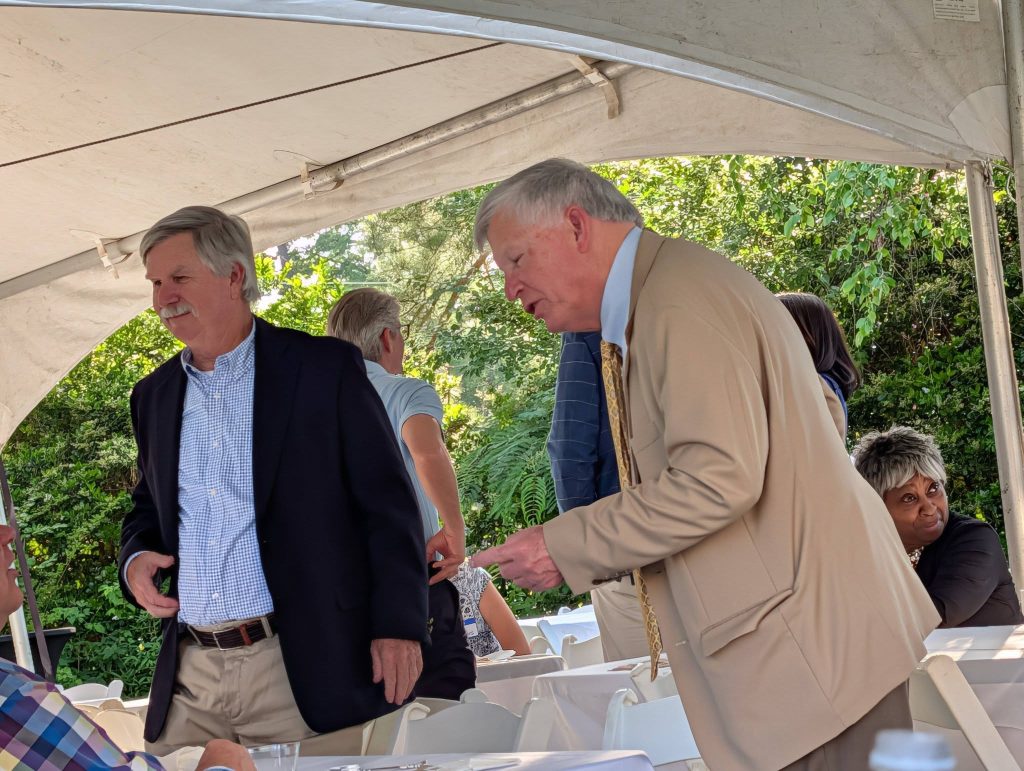 Two men in suits interact at an outdoor gathering under a tent, with other people seated nearby and greenery in the background.