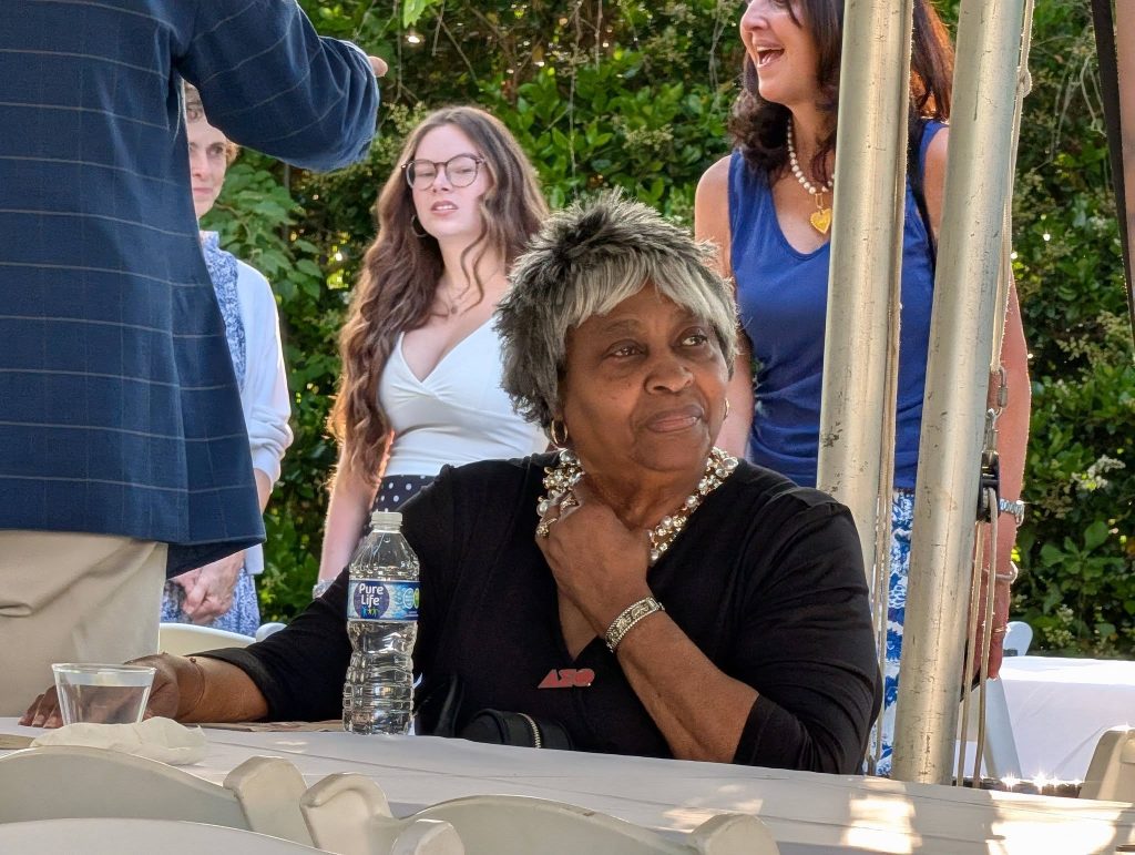 older woman sits at a table outdoors, surrounded by people standing and talking, with greenery in the background.