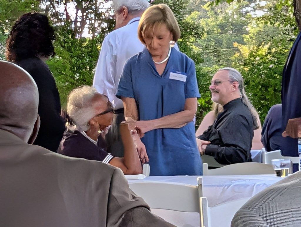 People gathered at outdoor event, some sitting at tables with white tablecloths, others standing and conversing, surrounded by greenery and sunlight in the background.