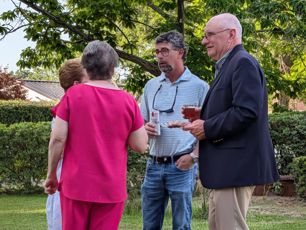 Four adults stand outside talking, holding drinks and food, surrounded by green trees and bushes on a sunny day.