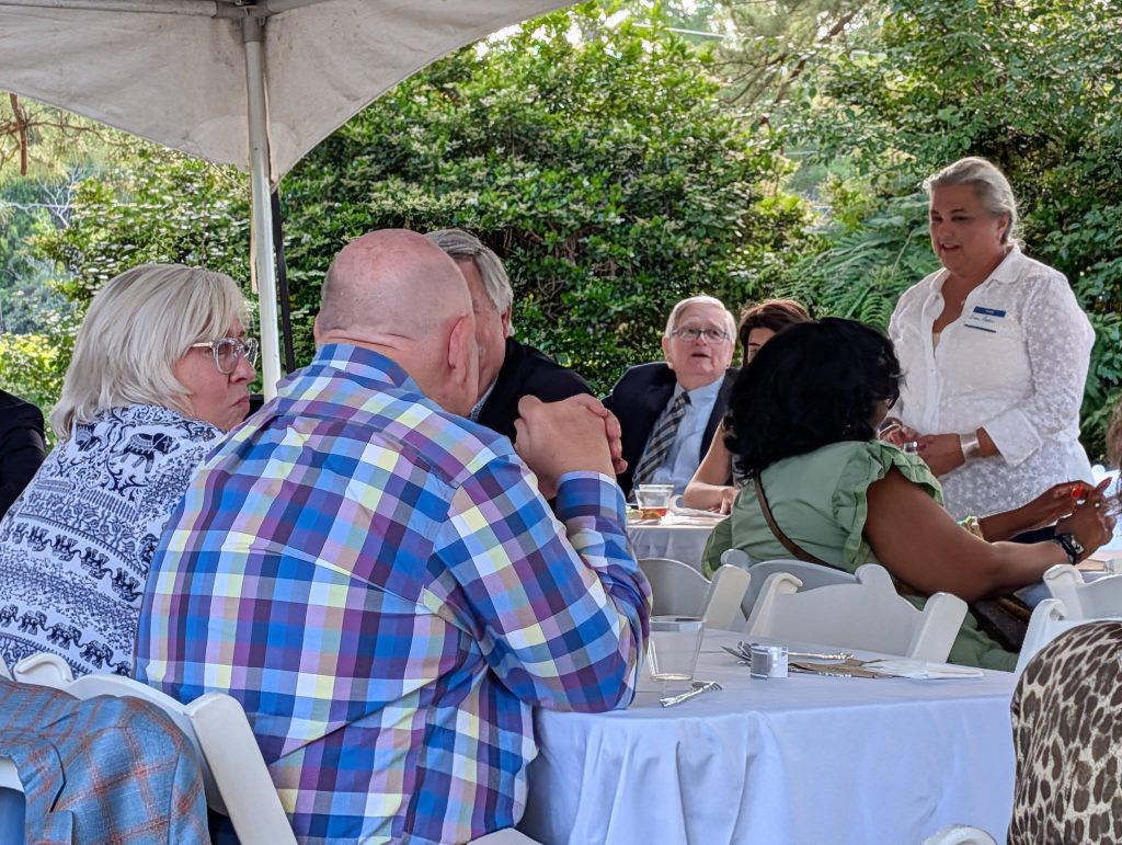 People sitting at round tables under a tent, engaged in conversation during an outdoor event with greenery in the background.