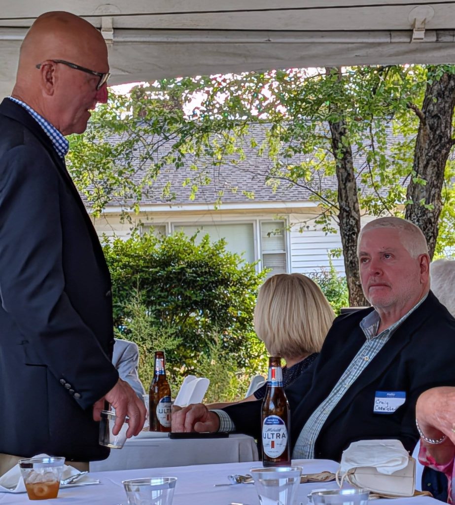 Three adults in formal attire sitting and talking at an outdoor event under a tent, with drinks and food on a white tablecloth.