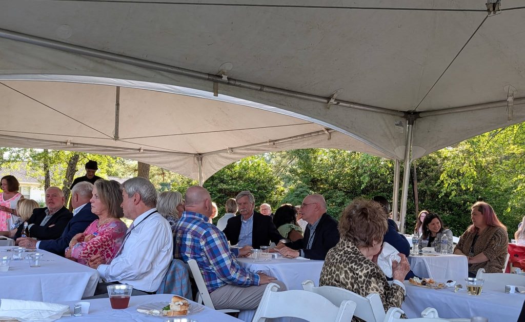 People seated at round tables under a white tent, enjoying food and drinks at an outdoor gathering with trees in the background.