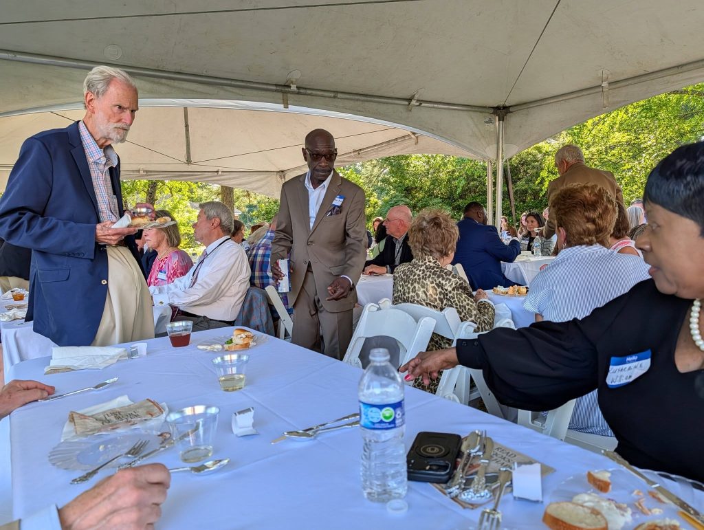 People gathered under a tent for a formal outdoor event, seated at tables with food and drinks, engaging in conversation and socializing.