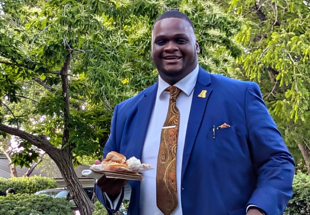 Man in a blue suit and patterned tie holding a plate of food, smiling outdoors with green trees in the background.