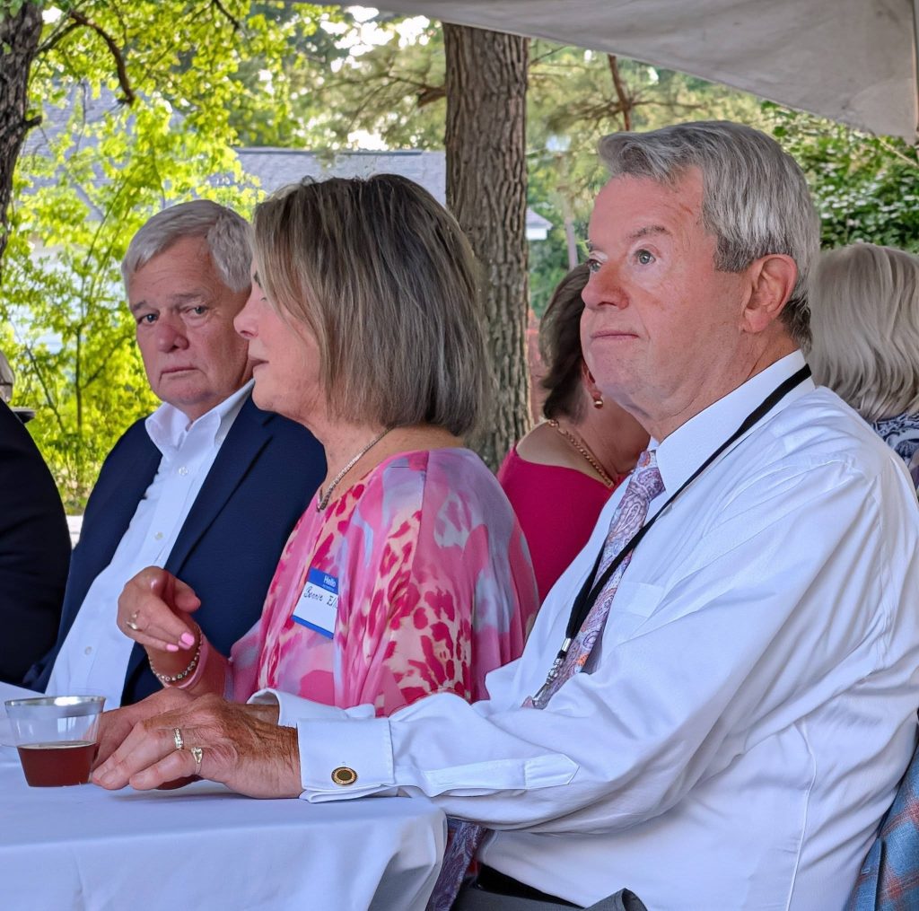 Three older adults sit at a table outdoors, dressed in formal attire, under a canopy, appearing focused and engaged in conversation.