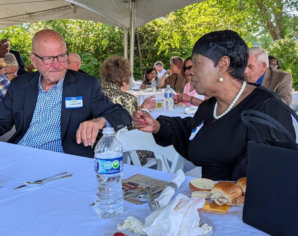 Two people sit at a table outdoors, talking during a gathering, with food and drinks on the table and other attendees in the background.