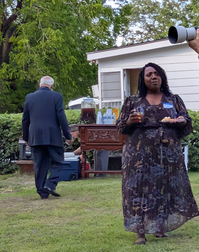 Woman holding food and drink at outdoor gathering; man in suit walks away. Greenery and a house are visible in the background.