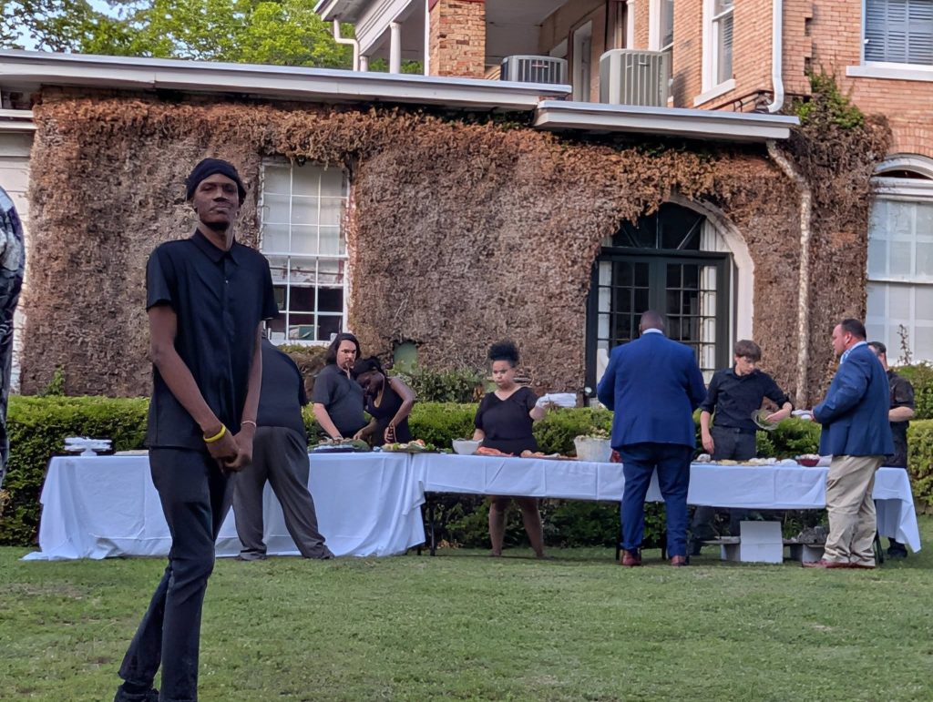 People gather around outdoor buffet tables on a lawn, with some serving food and others socializing in front of a building covered in dried vines.