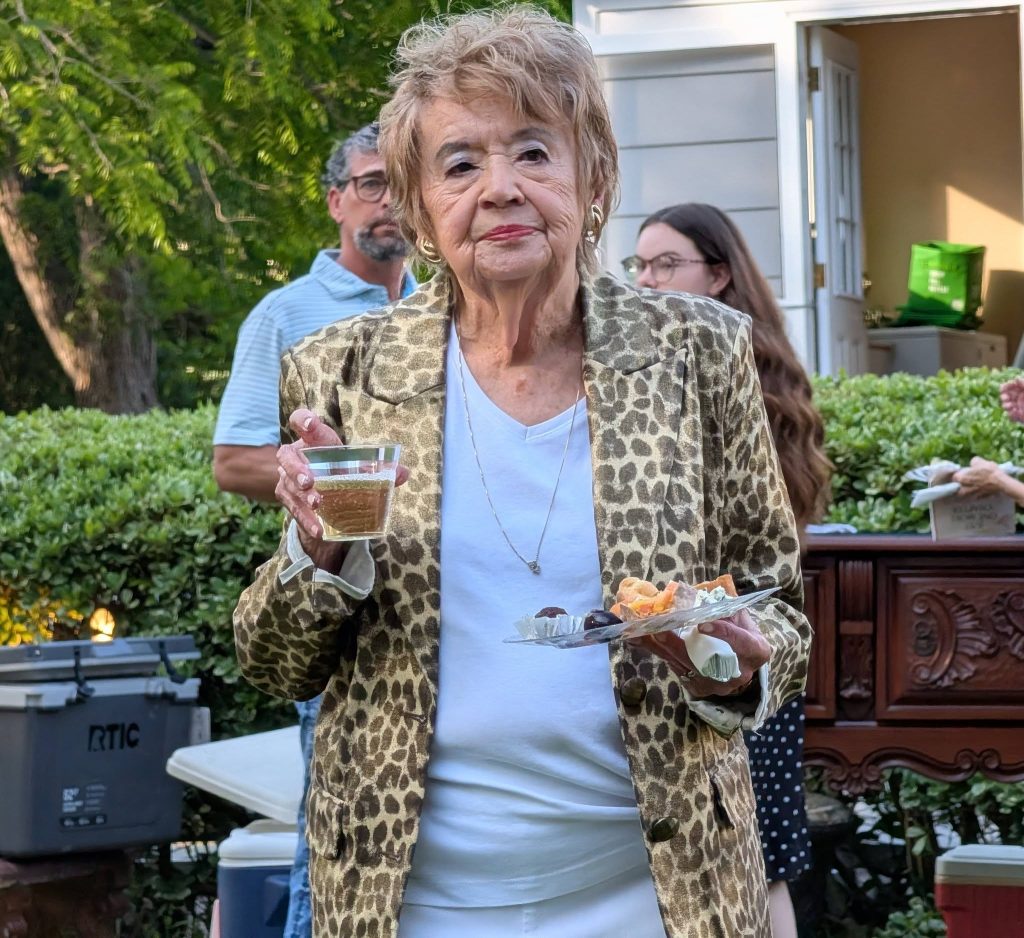 Elderly woman in a leopard print blazer holds a drink and a plate of snacks at an outdoor gathering, with people and greenery in the background.