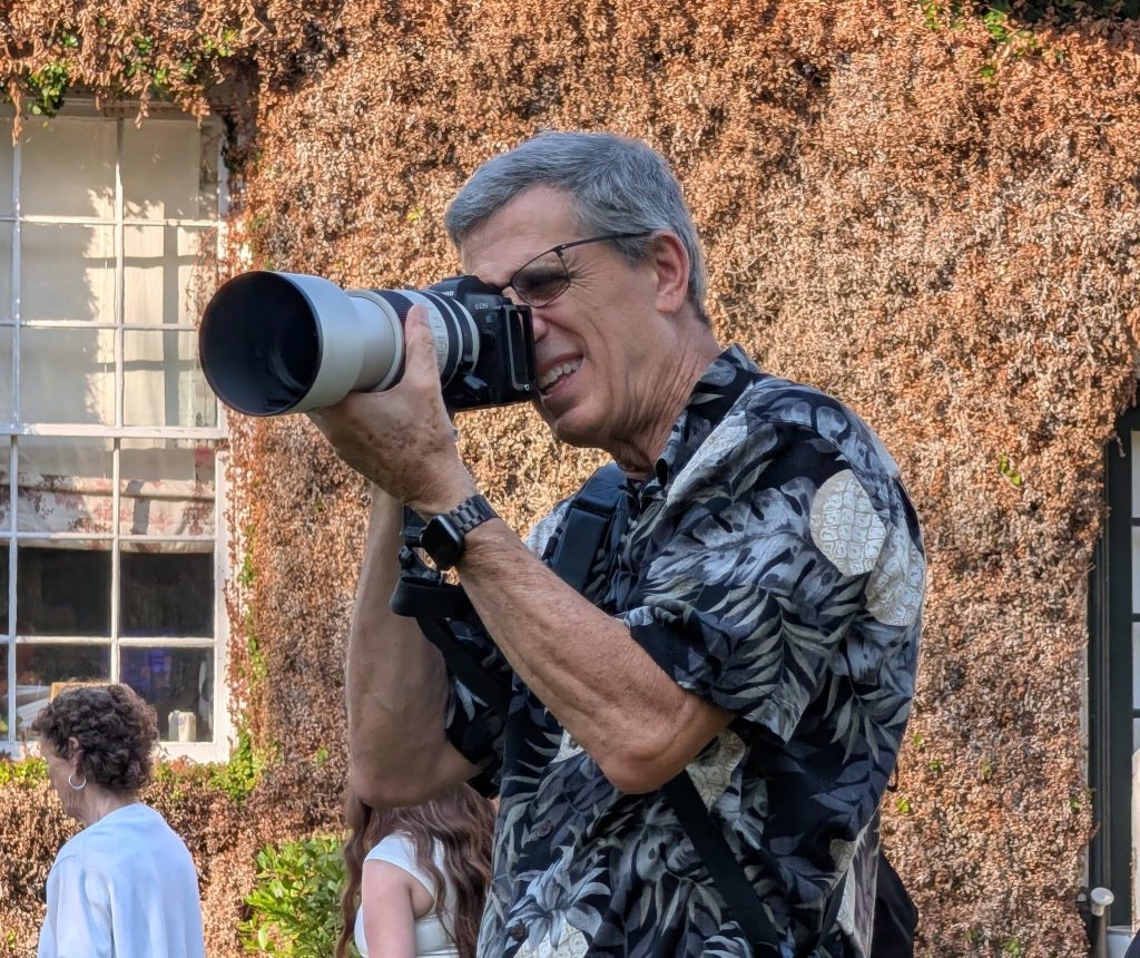 older man with gray hair and glasses is taking a photo with a large camera outdoors, wearing a patterned shirt.