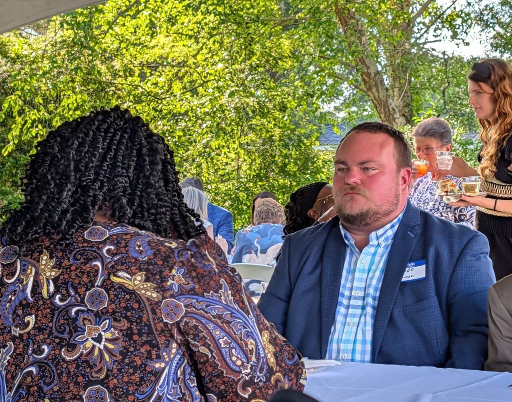 A man in a blue blazer sits at a table outdoors during a social gathering, facing a woman with curly hair, while others mingle in the background.