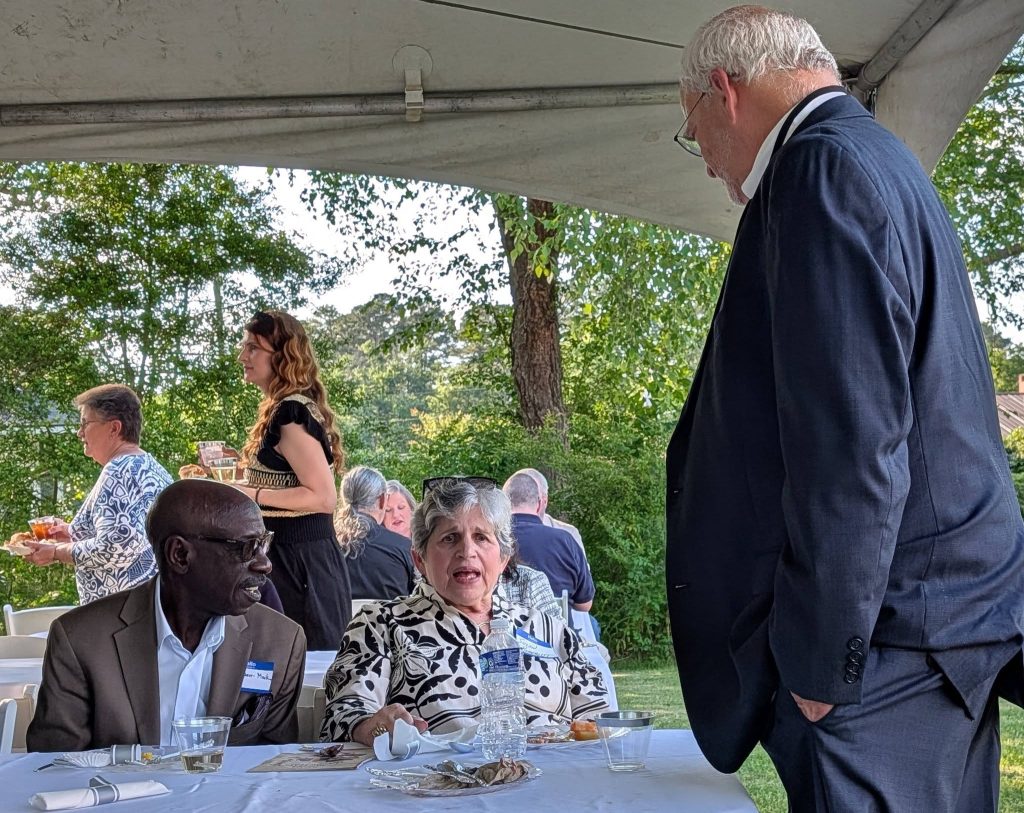 People gathered under a tent at an outdoor event, some sitting and talking, others standing with drinks, surrounded by trees and greenery.