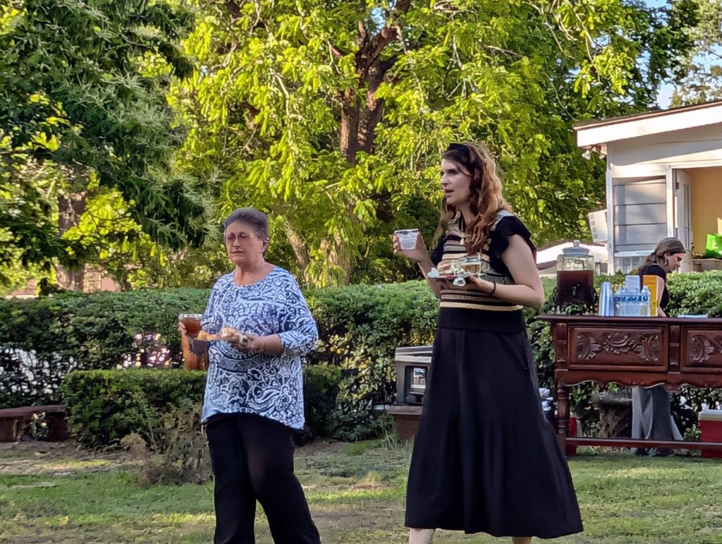 Two women holding drinks and food stand outdoors in a garden area, with a refreshment table and greenery visible in the background.