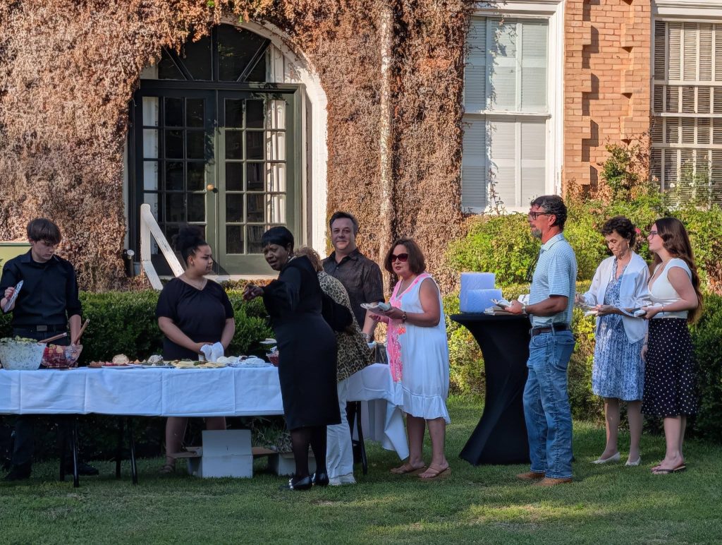 People lined up at an outdoor buffet table, serving and waiting for food, with a building covered in vines in the background on a sunny day.