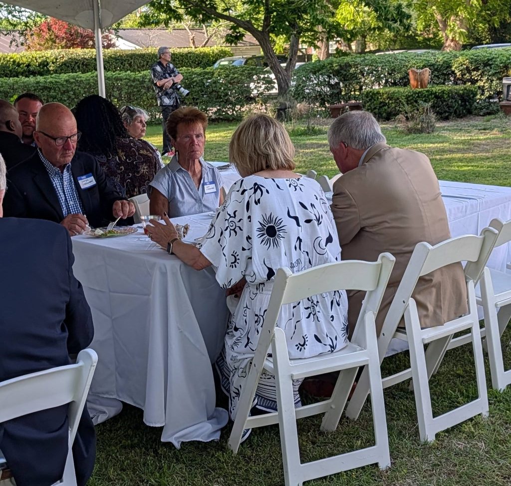 People sitting at a table outdoors under a tent, eating and talking during a social gathering, with greenery in the background.