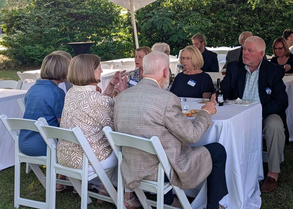 A group of people sit at a table outdoors, talking and eating during a formal event with white tablecloths and chairs.