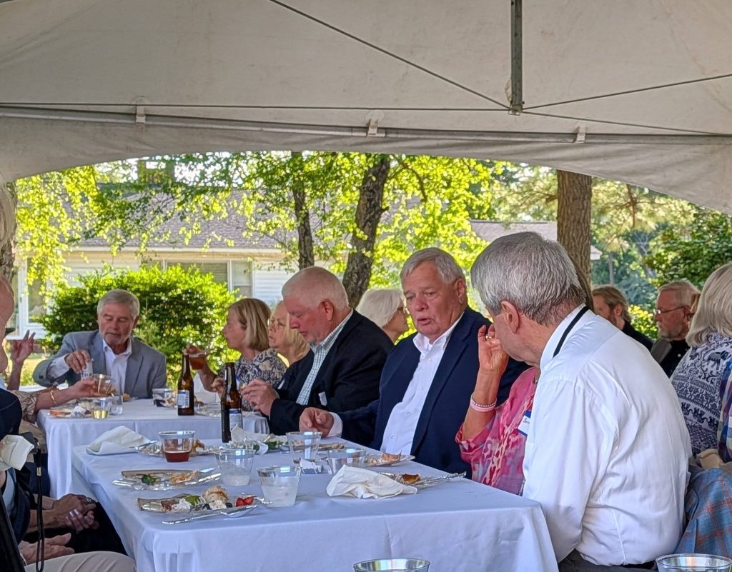 A group of older adults sitting at outdoor tables with white tablecloths, eating and drinking under a tent at a daytime gathering.
