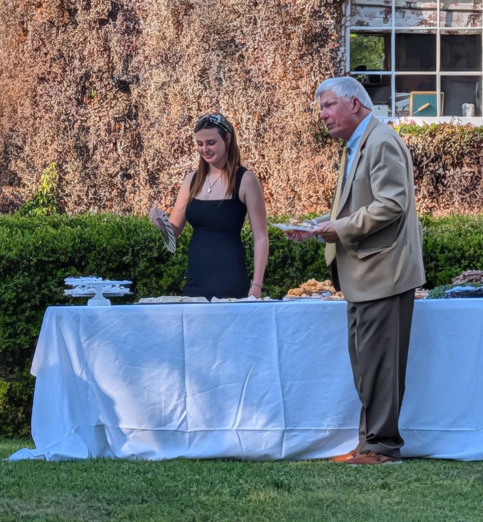 A young woman and an older man stand at an outdoor table covered with a white tablecloth, serving food on a sunny day.