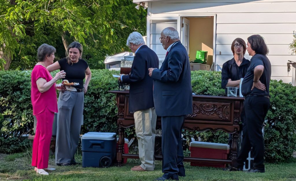 Six people stand and converse outside near a table, surrounded by greenery and a white house in the background. Some hold food and drinks.