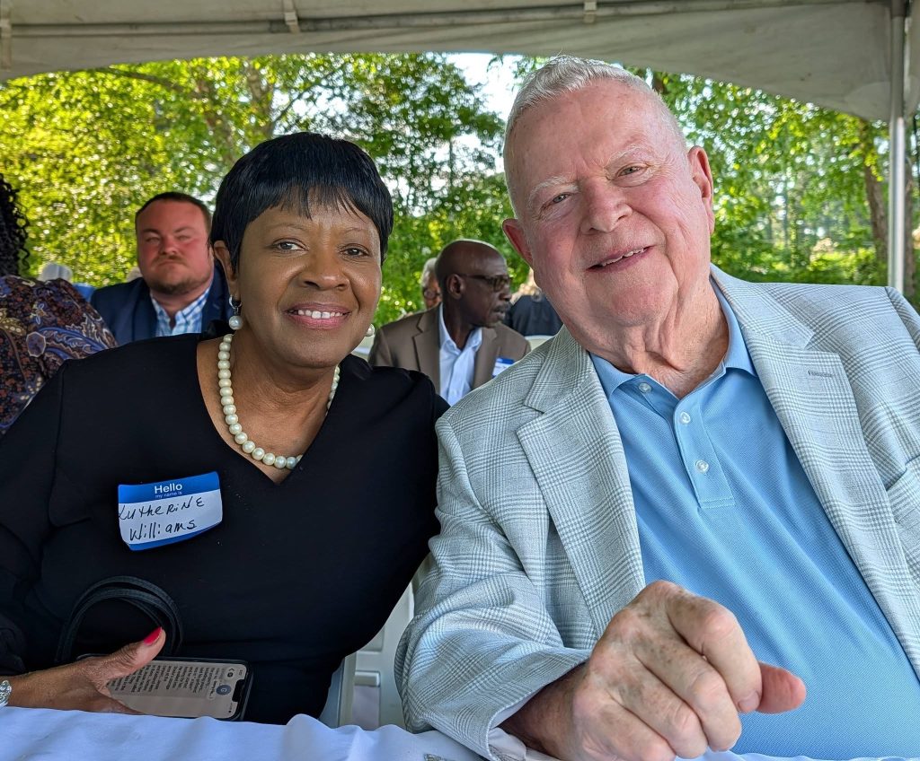 Two people smiling and sitting together at an outdoor event, surrounded by other attendees under a canopy, with green trees visible in the background.