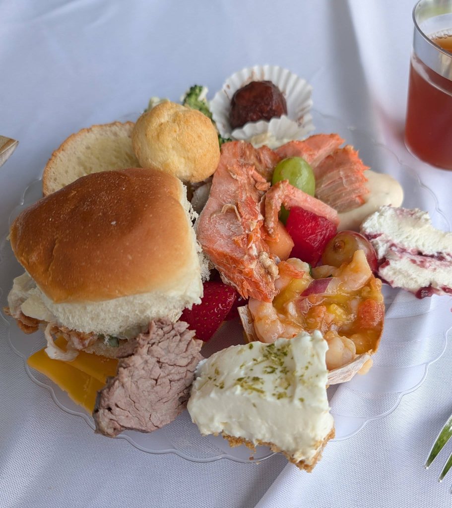 A plate with assorted foods including bread, salmon, shrimp, roast beef, fruit, cheesecake, a dessert ball, and a drink on a white tablecloth.