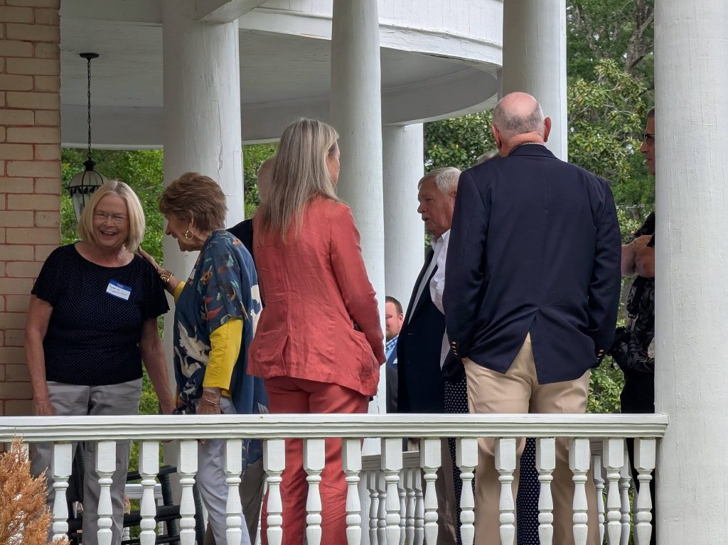 A group of adults socialize and converse on a porch with white columns and railing, surrounded by greenery on a cloudy day.