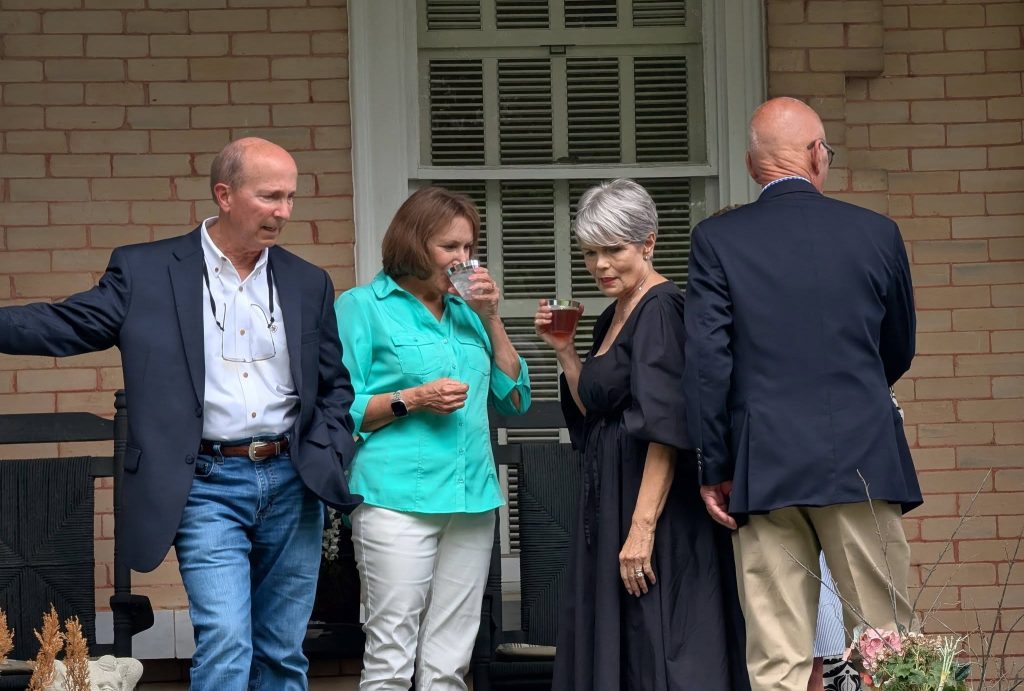 Four adults conversing on a porch, two women holding drinks. The men wear jackets, one with jeans, and the women wear teal and black outfits.