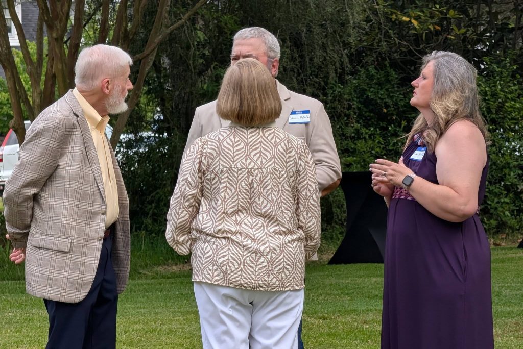 Four adults standing on grass, engaged in conversation at an outdoor event, wearing casual and business-casual attire with nametags visible, surrounded by trees and greenery.