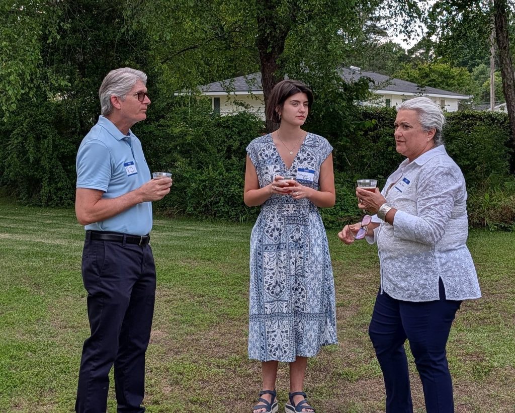 Three people with name tags stand outside on grass, holding drinks and talking, with trees and a house in the background.