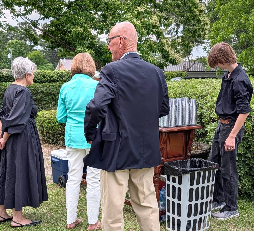 Four people stand outdoors near a table with stacked cups, a cooler, and a trash bin, surrounded by greenery and trees.