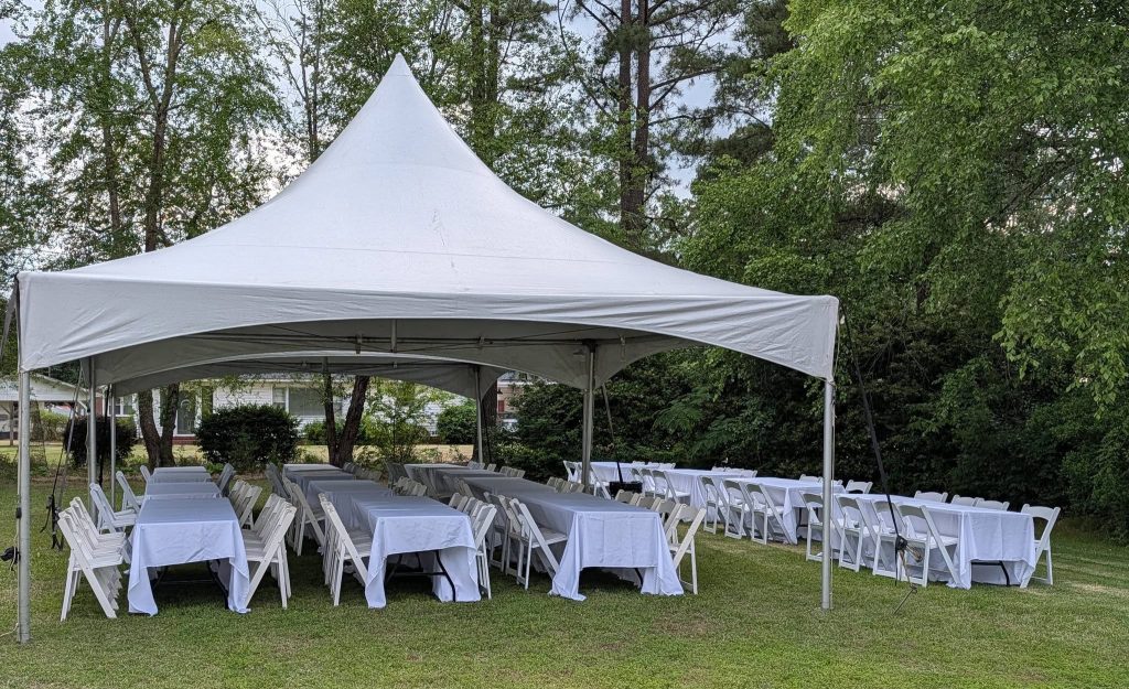 White tent set up outdoors with several tables and white chairs covered in white tablecloths, surrounded by green trees and grass under a cloudy sky.