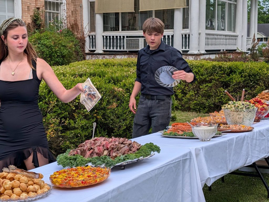 Two people at an outdoor buffet table with various dishes, including meats, salads, and pastries, in front of a large house with white columns.