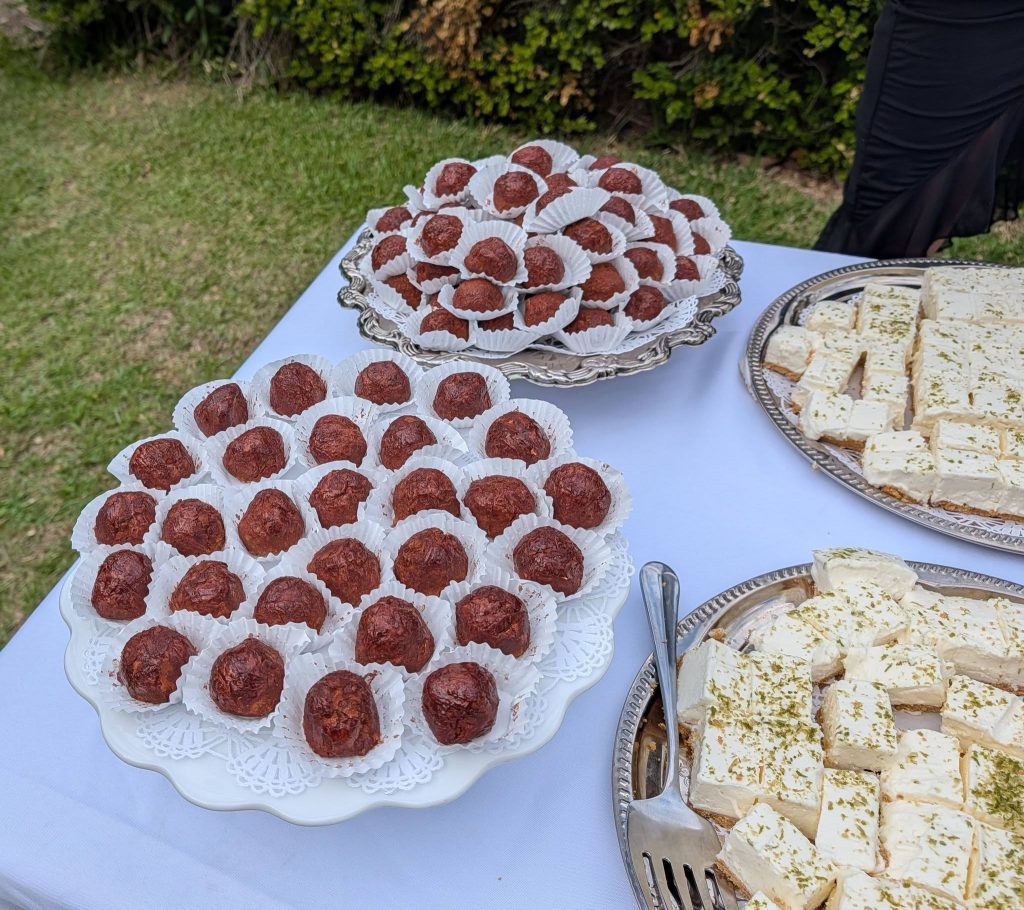 Assorted dessert trays with round chocolate truffles in paper cups and square pieces of white cake with green toppings on a white table outdoors.