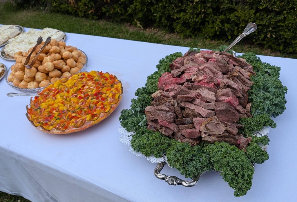 A buffet table with sliced roast beef on greens, cream puffs, a colorful fruit salad, and sandwiches on a white tablecloth outdoors.