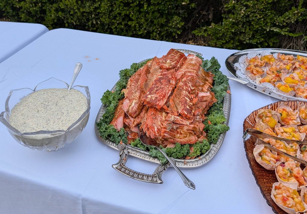 A buffet table with a platter of cooked fish, a bowl of creamy herb sauce, and trays of seafood appetizers in cups.