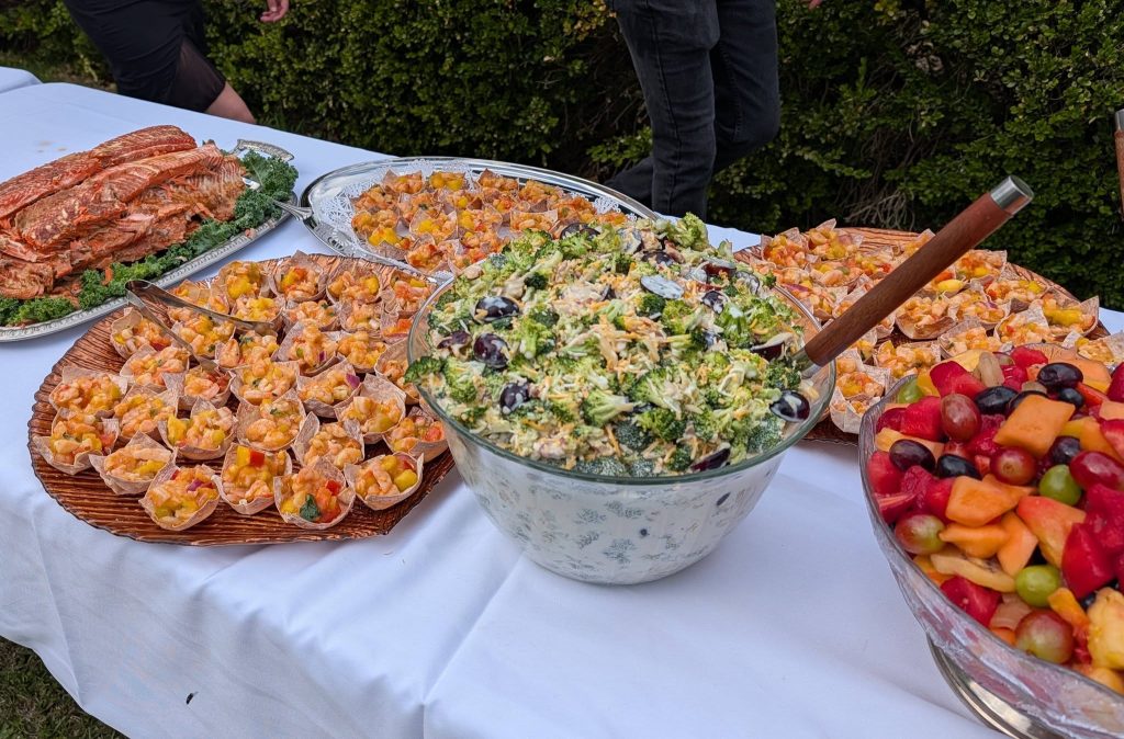 Buffet table with assorted foods including salmon, mini appetizers, broccoli salad with grapes, and a bowl of mixed fruit on a white tablecloth outdoors.