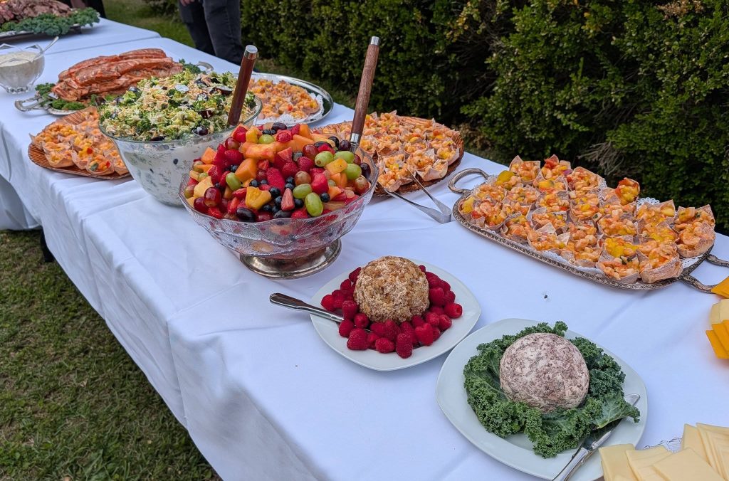 A buffet table outdoors with fruit salad, salads, cheese balls, assorted finger foods, and sliced meats arranged on white tablecloths.