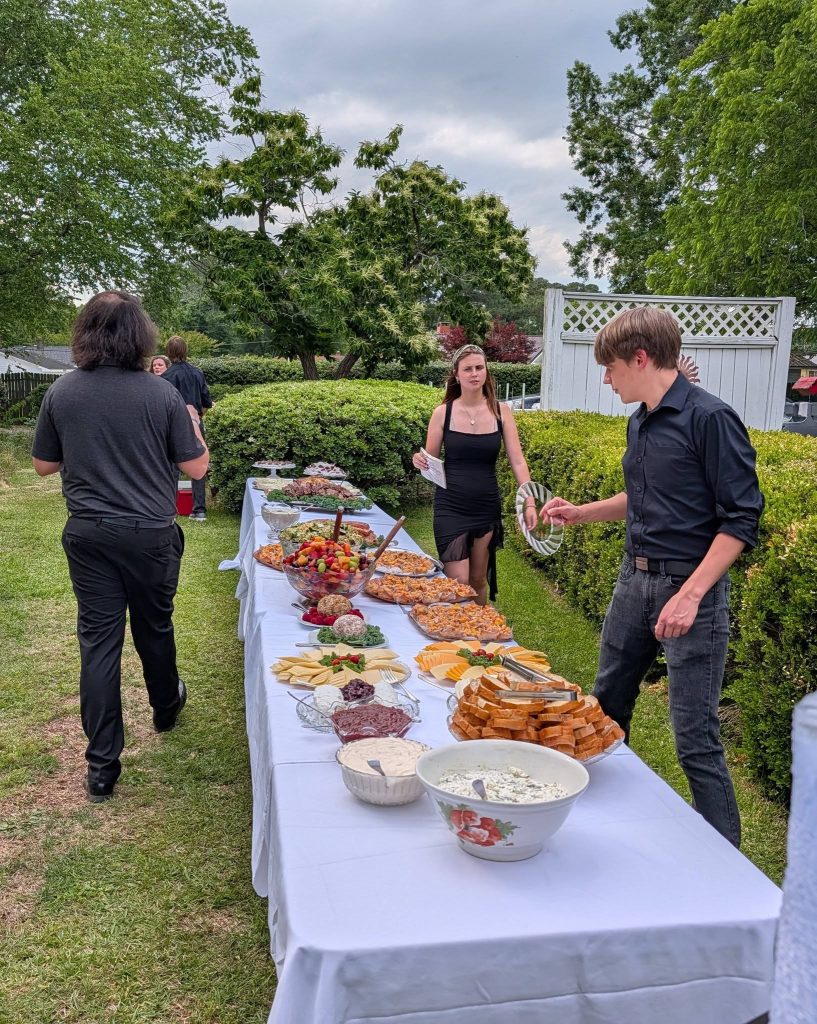 People serving themselves at an outdoor buffet table with assorted breads, cheeses, dips, and fruit under cloudy sky, surrounded by green trees and hedges.