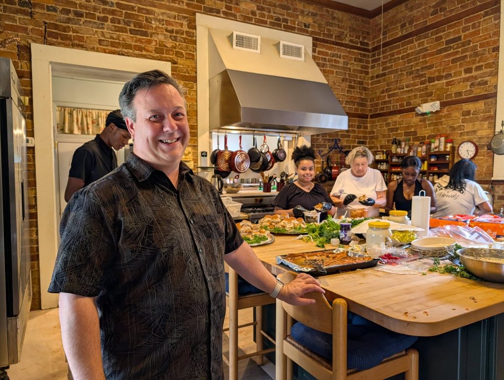 Smiling man stands in front of a busy kitchen island where several people prepare food, with various ingredients and dishes spread out on the wooden counter.