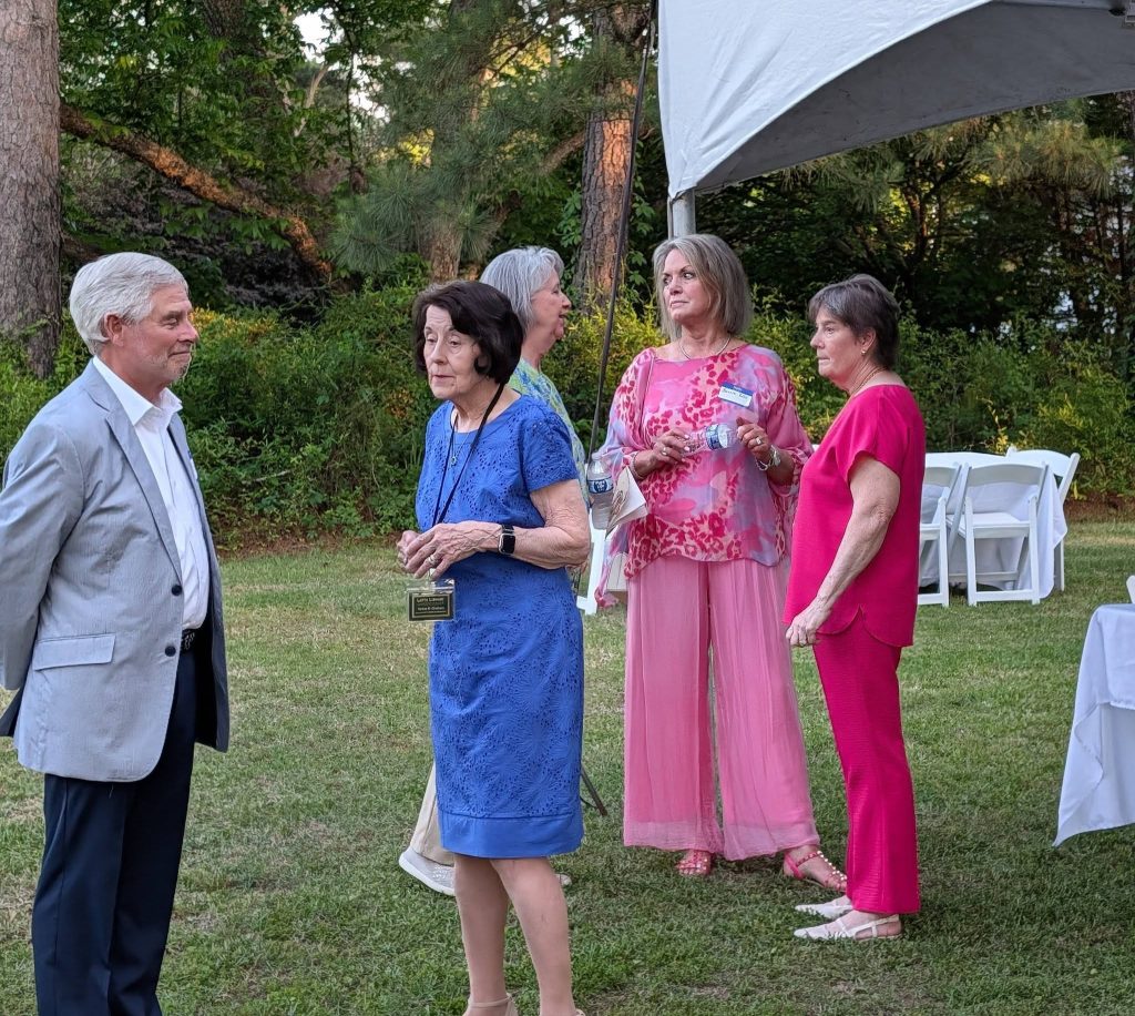 Five adults talk outdoors at a casual event. Four women wear colorful dresses and one man is in a gray suit. White chairs and trees are in the background.