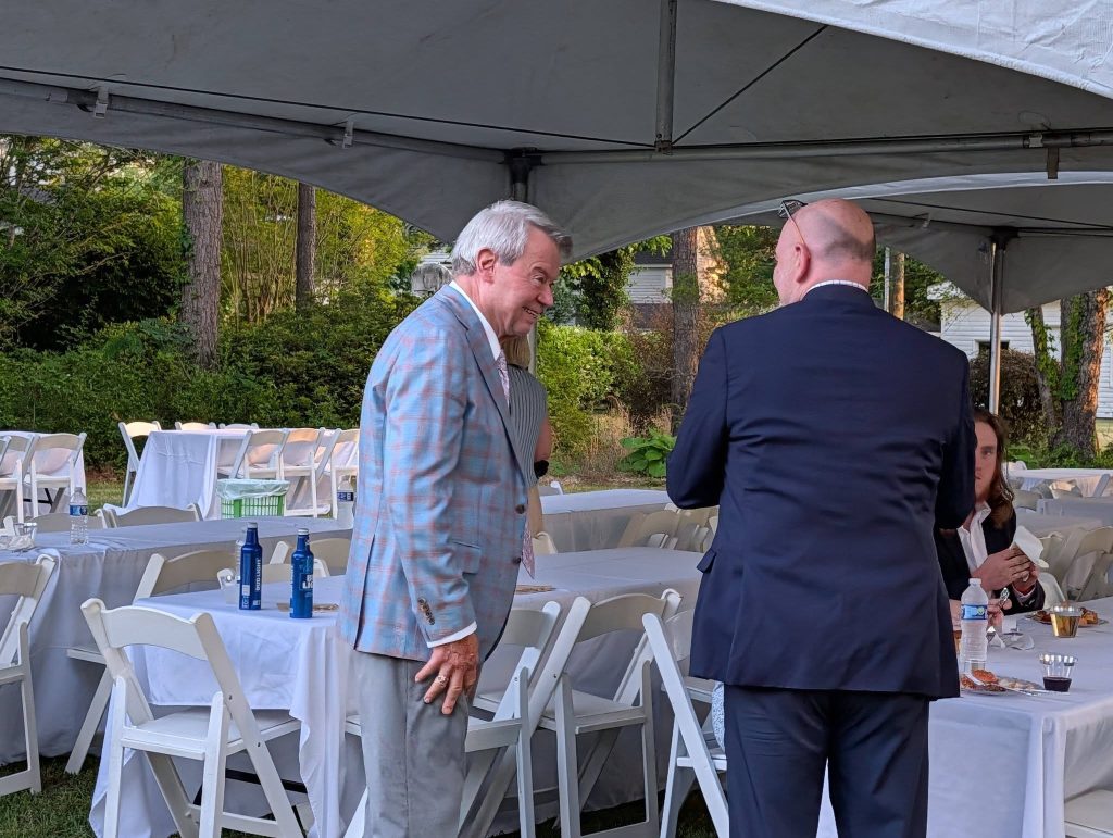 Two men in suits talking under a large outdoor tent with white chairs and tables. Some people are seated and eating in the background.