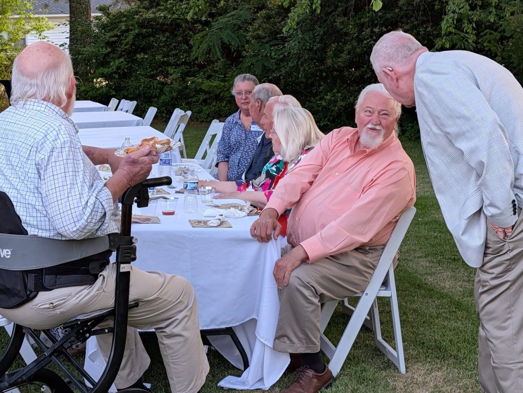 A group of elderly people sitting around a table outdoors, eating and talking, with white chairs and tablecloths, surrounded by green trees.