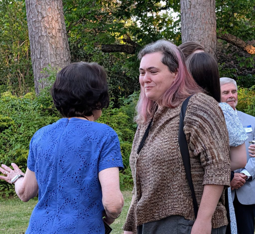 Two women are talking at an outdoor gathering, with trees and greenery in the background. Other people are visible in the scene.