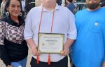 Young man holding a certificate of completion, standing outside with two adults, possibly family, during a graduation or school event.