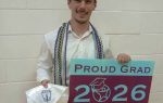 A smiling young man wearing honor cords holds a graduation cap and a sign reading "Proud Grad 2026 South Carolina Connections Academy."