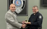 Two men shake hands as one presents a small trophy to the other, standing in front of a City of Dillon Police Department emblem.