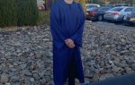 A person in a blue graduation gown and cap stands outside on a sidewalk near a rocky area and parked cars.