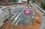 Steel building framework under construction, with a red flag displaying a large "S" and bulldog logo. Construction vehicles and materials are visible on the dirt ground.